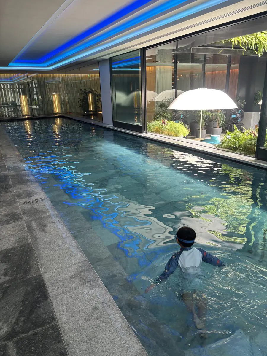 W大阪4階の屋内プールWET。細長いプールの天井に青いネオンが走り、水面に映り込んでいる。右手のガラス越しに屋外テラスのパラソルと植栽が見える