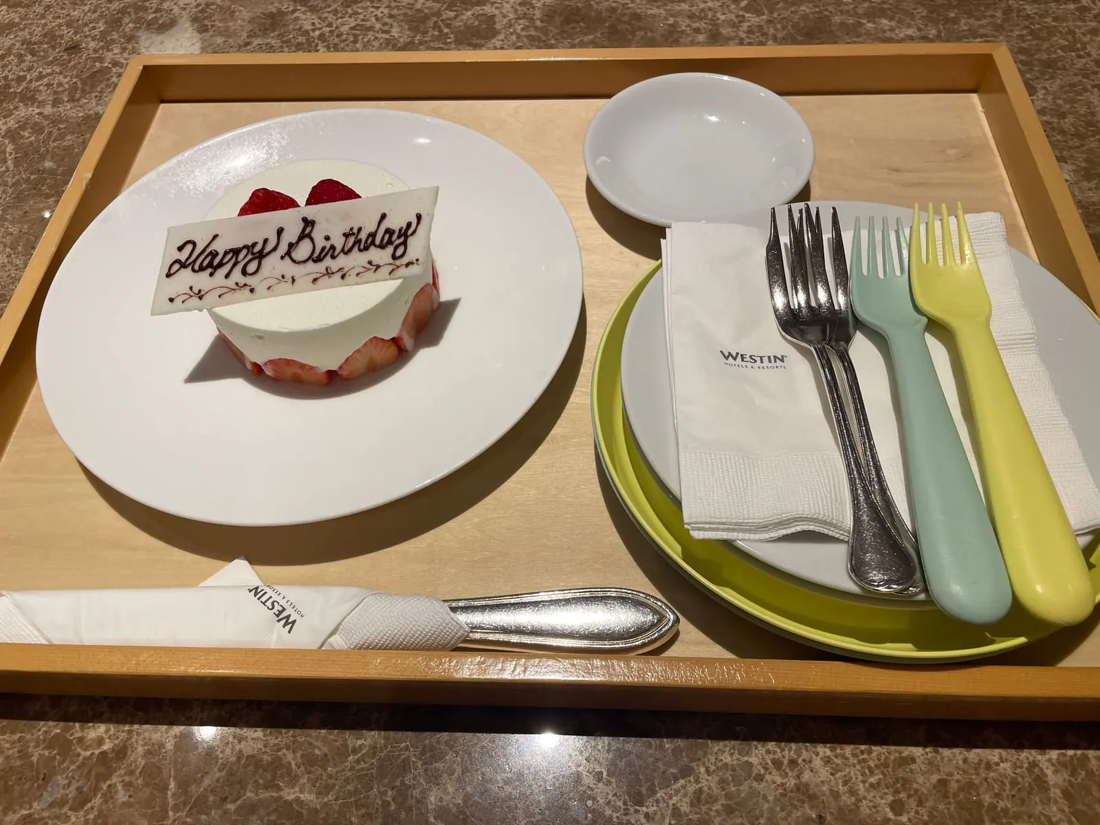 トレイに載ったバースデーケーキ。白いプレートにいちごのショートケーキと「Happy Birthday」のチョコプレートが添えられ、WESTINのロゴ入りナプキンとカトラリーが並んでいる